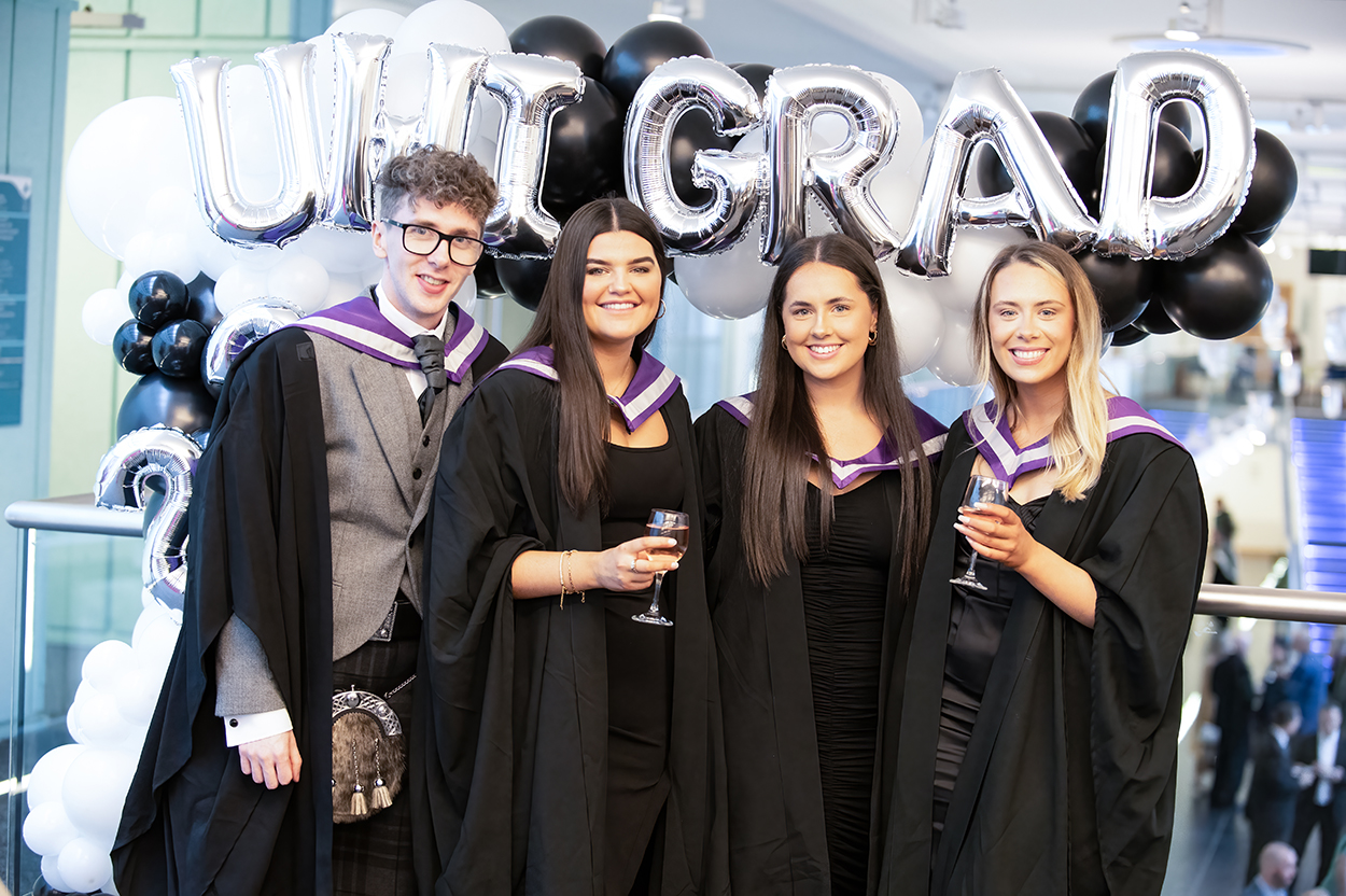 Students wearing robes, stood infront of balloons that say 'UHI GRAD'